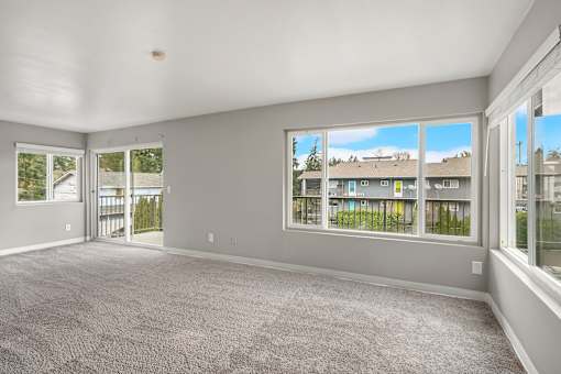 A spacious room with grey carpeting and large windows overlooking a residential area.